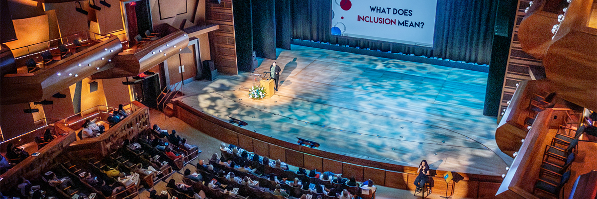 A presenter stands behind a podium under stage lights on the Musco Center stage, in front of a projector screen that reads, "What Does Inclusion Mean?" as an audience observes in the foreground.