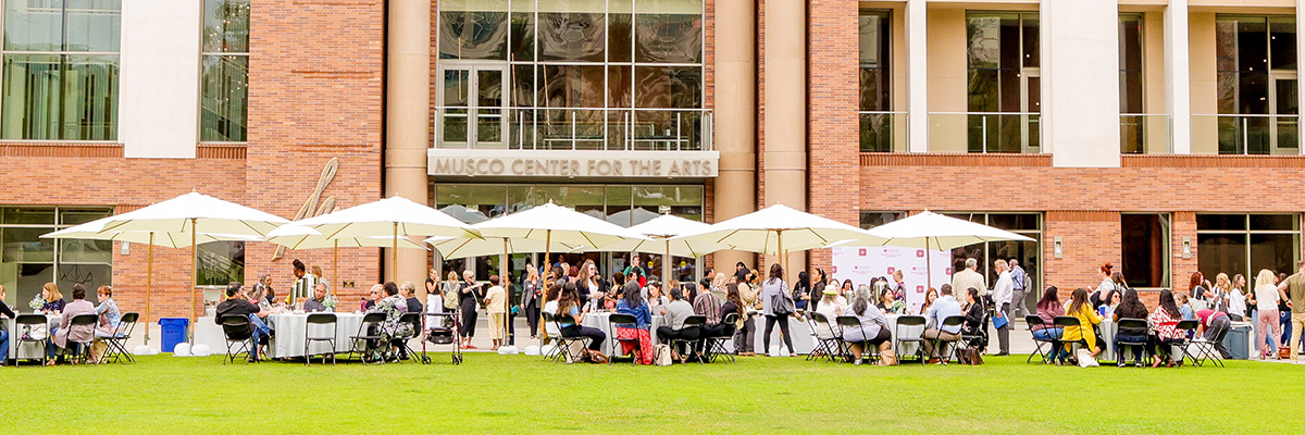Outdoor gathering at Musco Center for the Arts with people seated under white umbrellas.