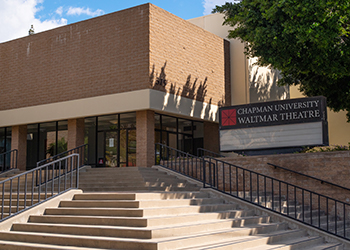 The front face of Waltmar Theatre. A large staircase leads up to the tall, red brick building with glass doors. To the right of the doors, the marquee sign rest beneath a tree.